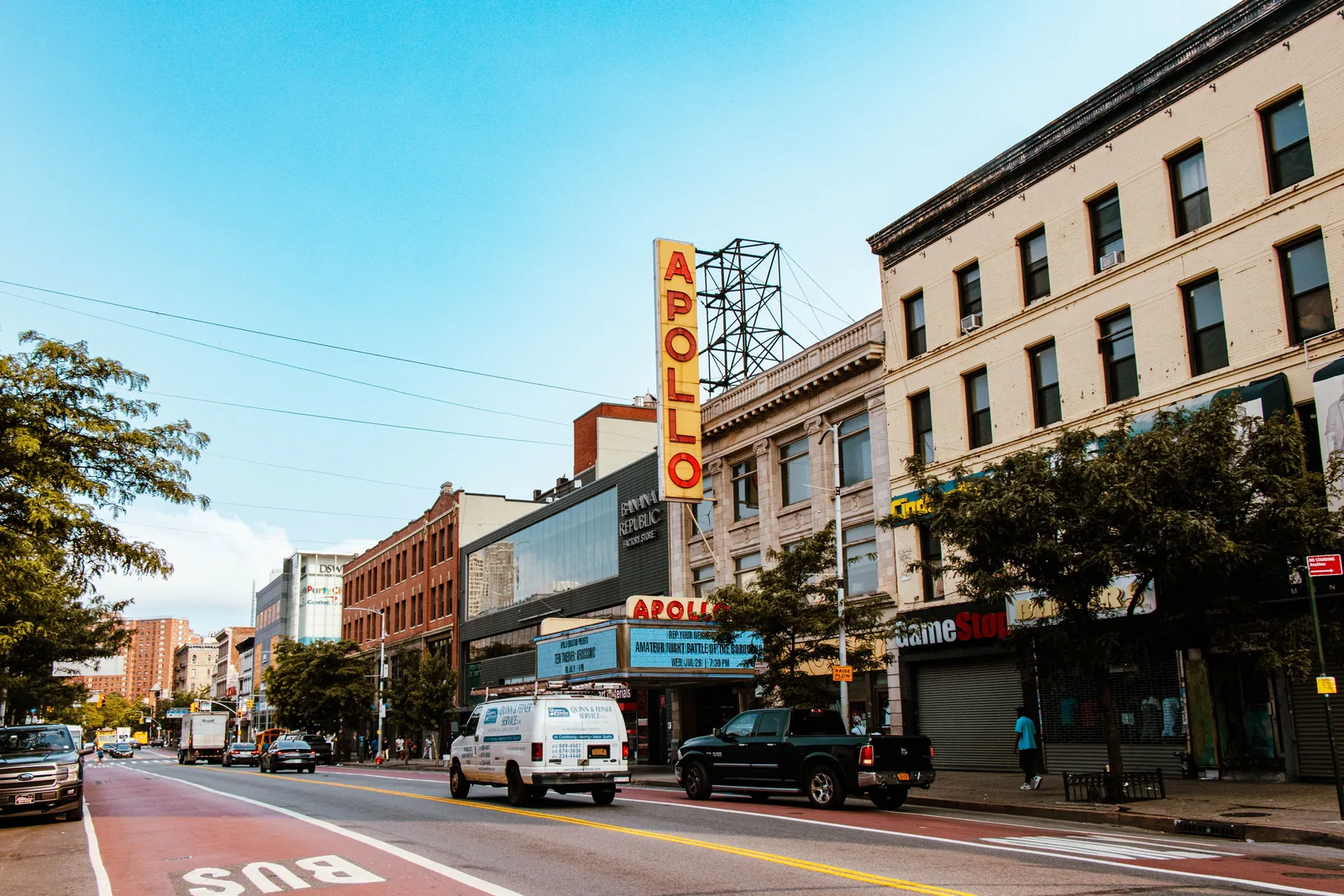 The Apollo Theater's tall vertical sign and marquee on 125th Street in Harlem, beside a red bus lane and brick buildings under a blue sky