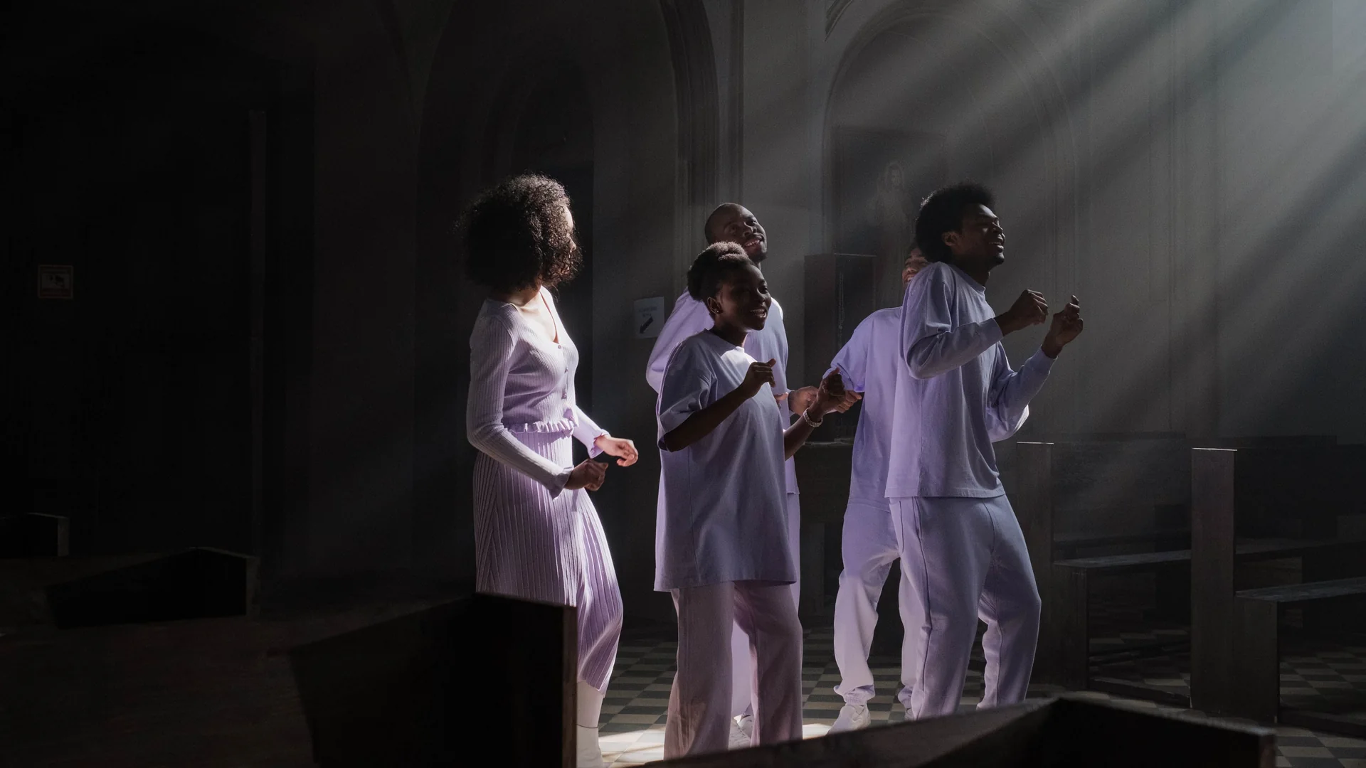A group of singers in pale lilac clothes sing and clap in a dim church while shafts of daylight fall through the air around them; a general photograph of gospel singing, not of this venue