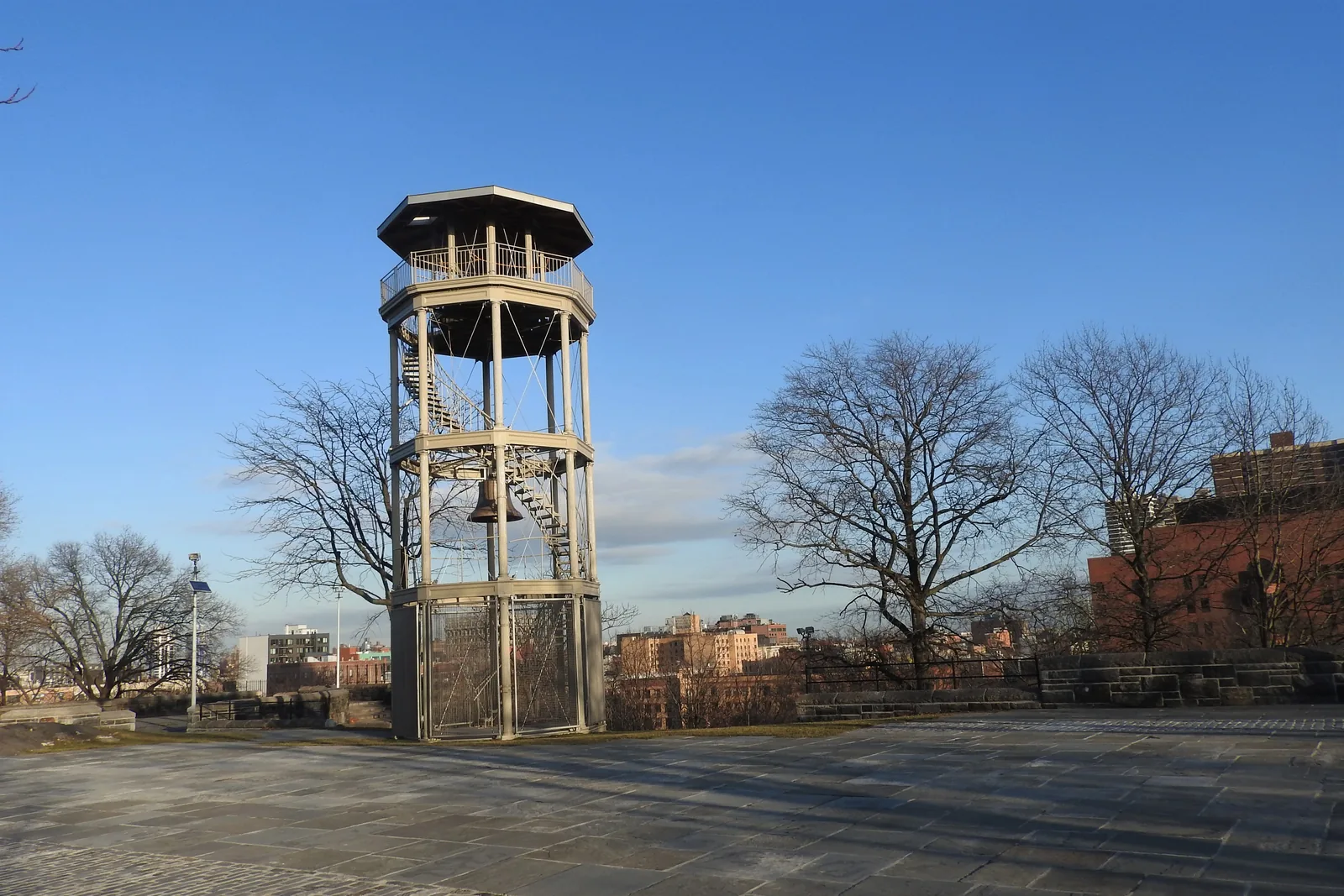 The octagonal cast-iron fire watchtower in Marcus Garvey Park, with its bell hanging inside, on a stone plaza under a clear blue sky